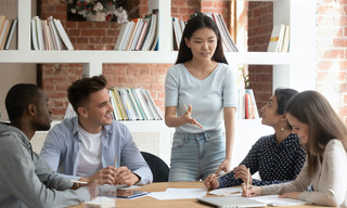 Smiling motivated asian female team leader holding meeting with diverse groupmates, explaining new project detail. African american, indian and caucasian concentrated students discussing assignment